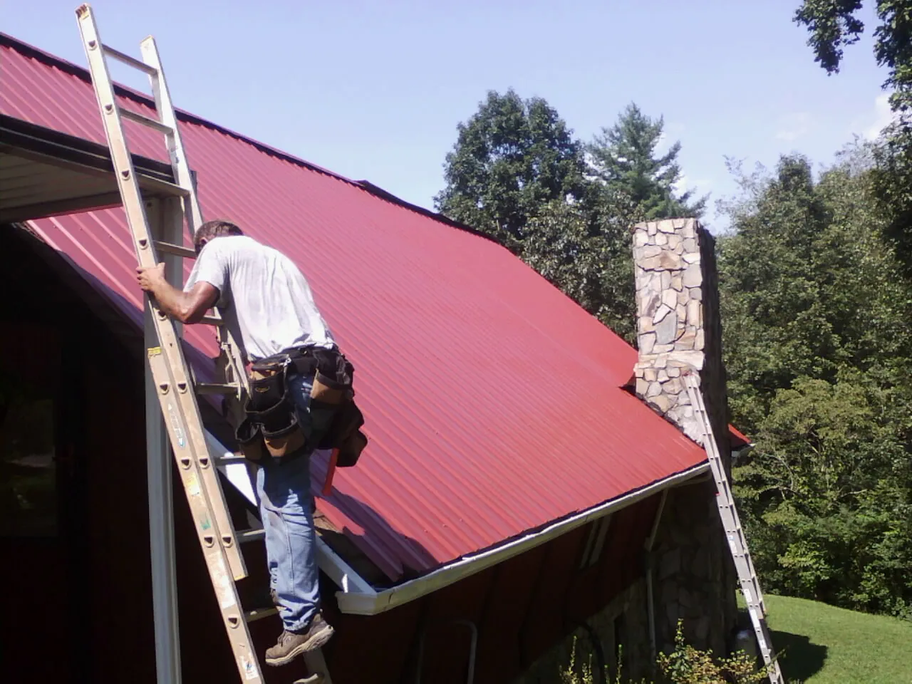 Roofer on a ladder beside a red metal roof and stone chimney — Soffit & Fascia Repair by Roofing Brothers in Perley